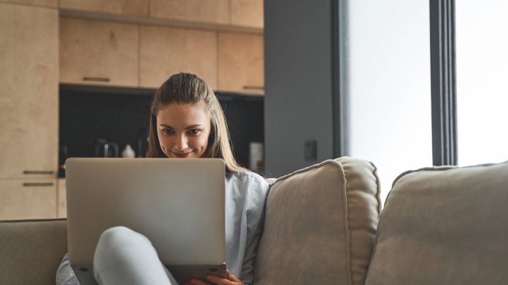 A young female with long brown hair smiles to herself as she sits on a sofa at home and reads an article on her laptop that answers the question, 'Is Payoneer safe?'