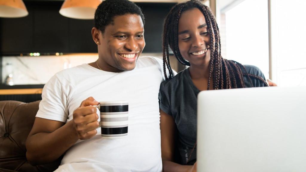 A man and woman sit next to each other and smile as they read an article on their laptop about sending automotive payments