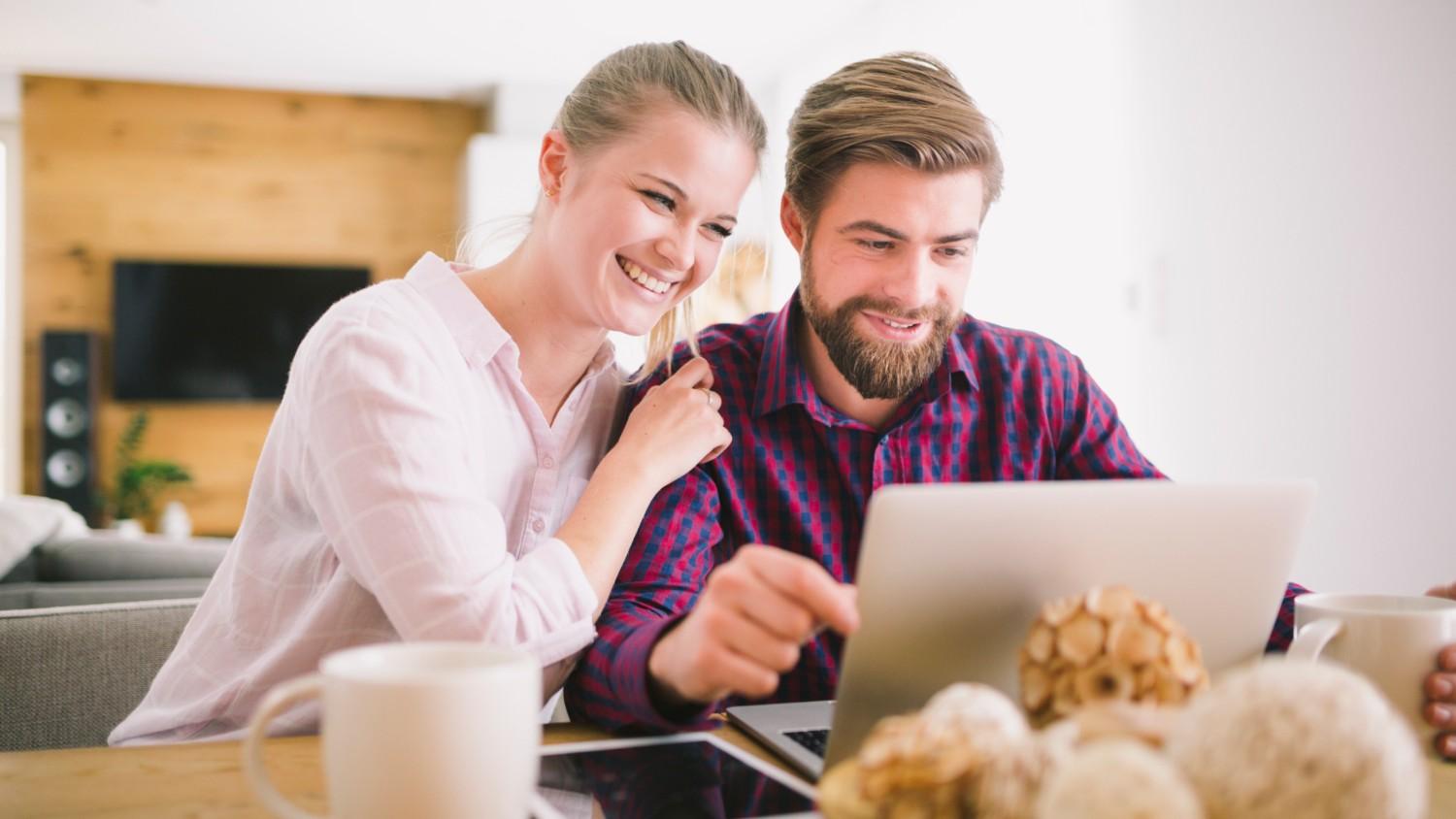 A young man and woman smile as they sit next to each other at a table and read the Revolut Account Review on their laptop