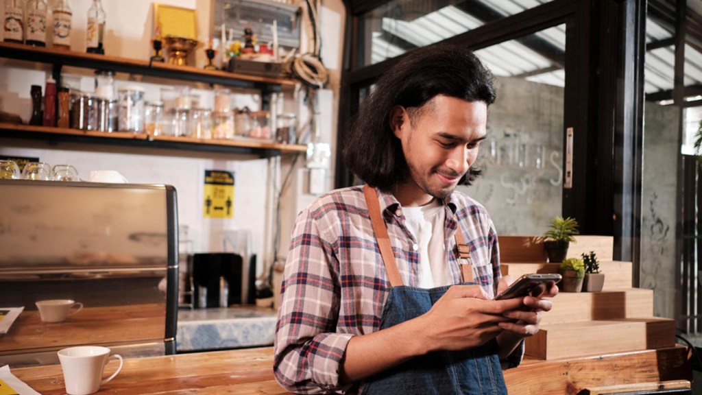 A young man in a checked shirt and apron stands behind a desk, reading an article on his phone about digital payment platforms