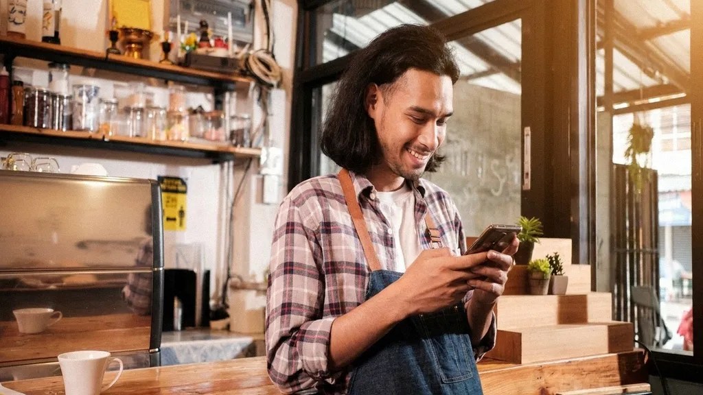 A young man in a checked shirt and apron stands behind a desk, reading an article on his phone about digital payment platforms
