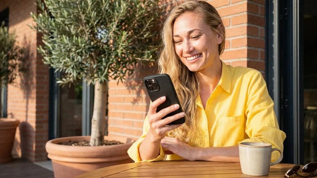 A young female with long blonde hair wears a yellow shirt and smiles as she sits outside next to a bush and holds her phone, reading an article about how to become sole trader in the UK