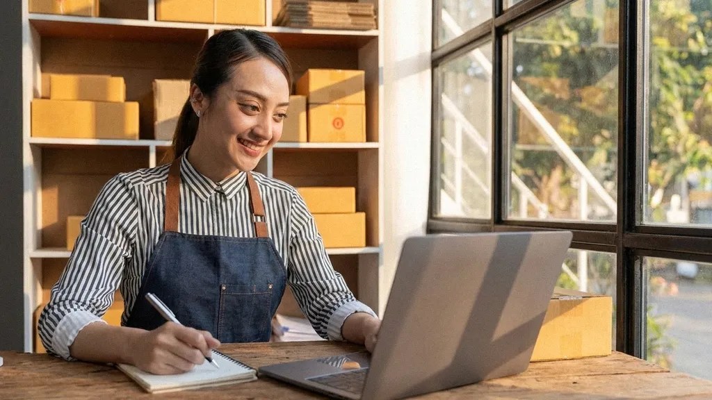 A young brunette female in a uniform sits at her desk and writes as she looks at her laptop to read an article about how to open business bank account in the UK
