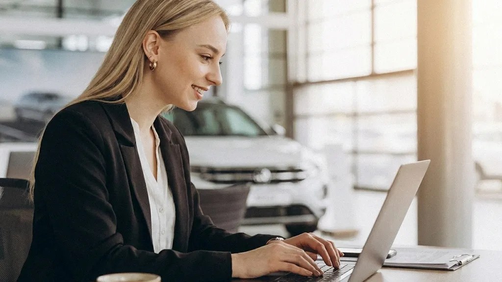 A young blonde female in a black blazer faces the camera as she sits at a desk and types on her laptop about how to manage business expenses