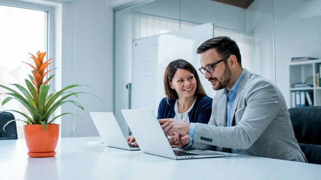 A young brunette female and a young man with glasses smile as they sit next to each other at a desk, typing on their laptops while discussing how to manage business expenses