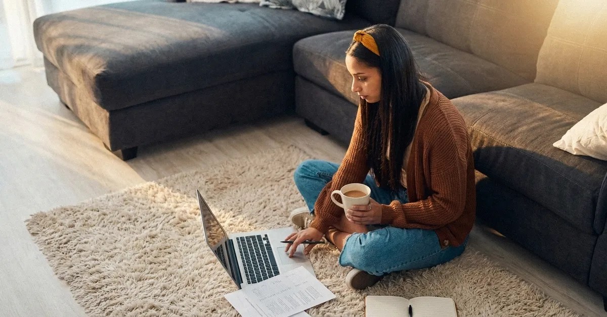 A girl with dark hair sits on the floor in front of her sofa, reading an article on her laptop about the cost of living in the UK