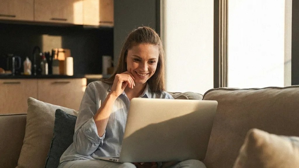 A young female with long brown hair smiles to herself as she sits on a sofa at home and reads an article on her laptop that answers the question, 'Is Payoneer safe?'