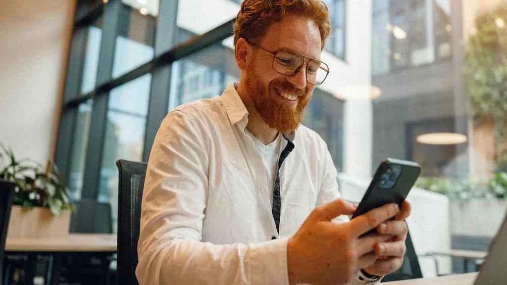 A bearded man wearing glasses and a white shirt smiles to himself as he checks his phone to ask the question 'Is Revolut Business safe?'