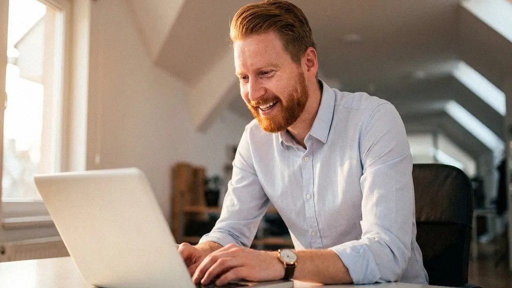 A young man with a beard wears a light shirt and sits at his office desk with his laptop while looking at the camera and smiling as he considers, 'Is Skrill Business safe?'