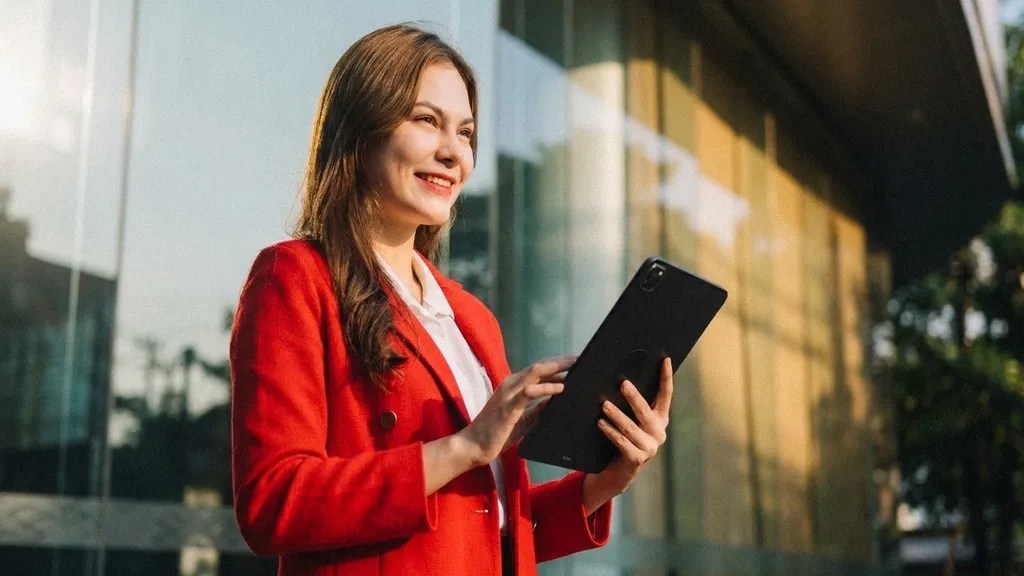 A young brunette female in a red suit  stands outside and smiles as she reads an article on her ipad about 'Is Wise Business safe?'