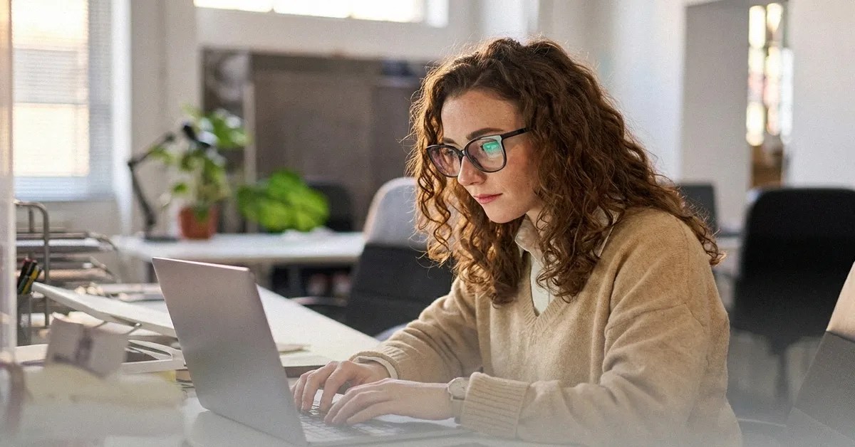 A young woman with brown curly hair and glasses sits at a table and reads an article about the best Revolut alternatives on her laptop