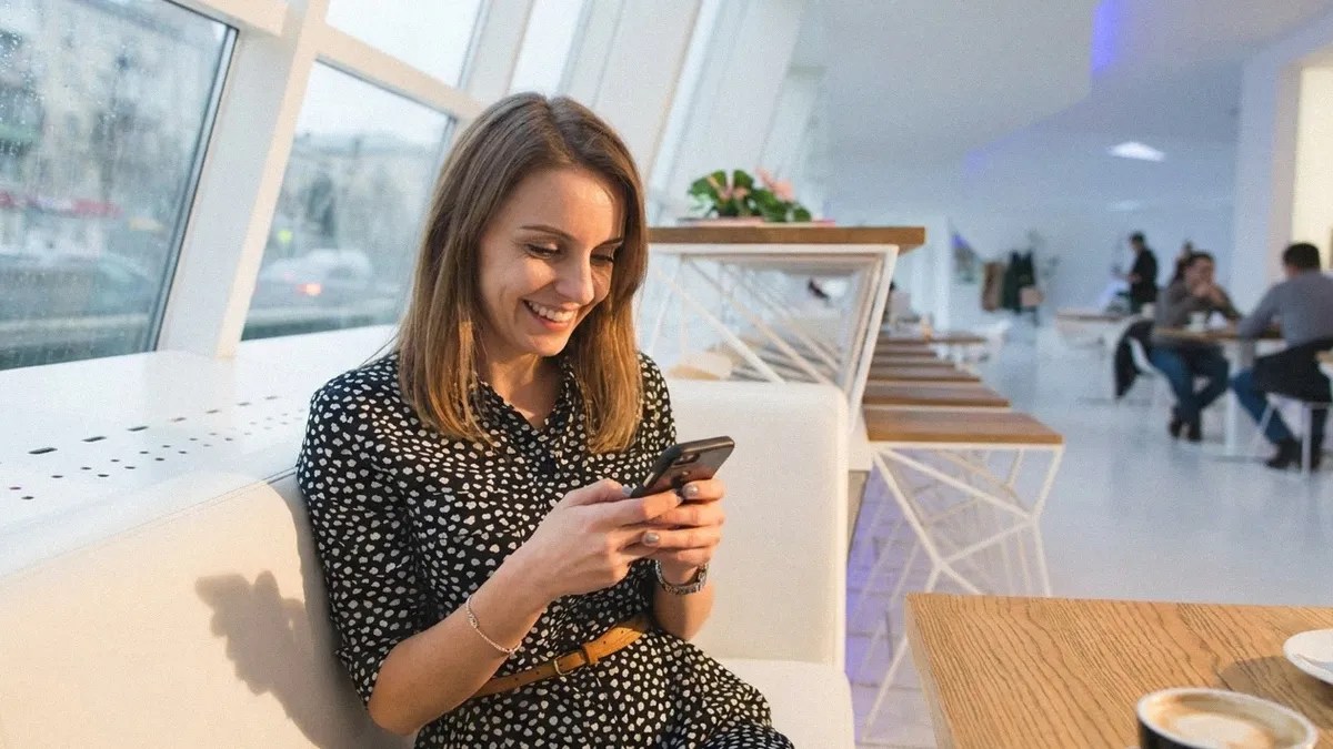 A young brunette female in a patterned shirt sits on the sofa and reads an article on her phone about how to pay international contractors