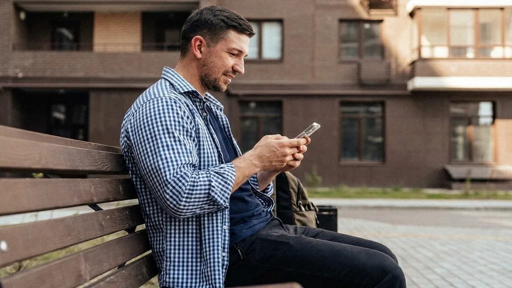 A young man in a blue check shirt looks ahead as he sits on a bench outside and reads about the Revolut Business Account on his mobile phone 