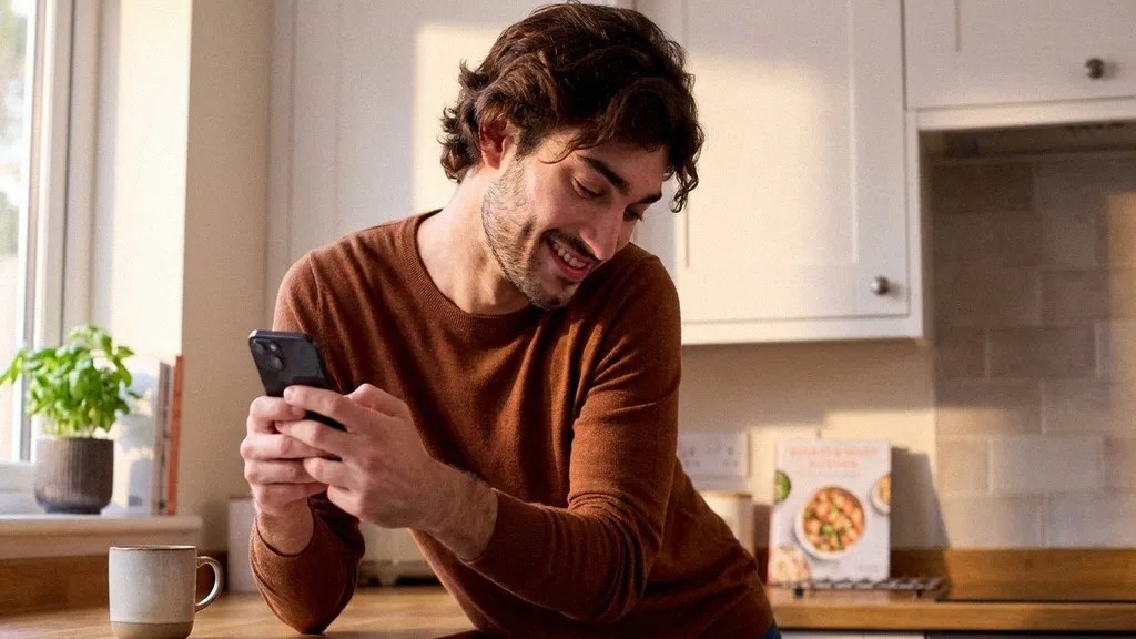 A young man smiles to himself as he stands in his kitchen reading the TransferGo vs Revolut Business comparison article on his mobile phone
