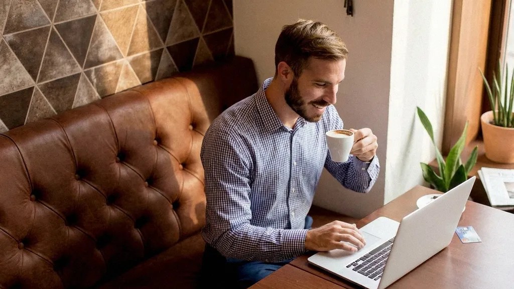 A young bearded male in a checked shirt sits at a table in a restaurant, drinking coffee and reading an article on his laptop about TransferGo vs Luminor Business