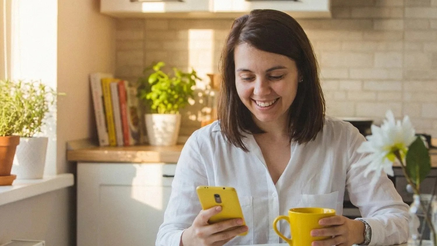 A smiling brunette female sits at the table in her kitchen, reading an article on her phone about the Wise business account